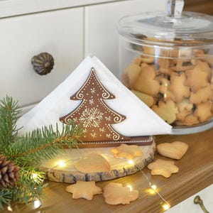 Pode incluir: Uma cena festiva de Natal com um porta-guardanapos castanho em forma de árvore, guardanapos brancos e biscoitos em forma de coração e estrela numa fatia de madeira. Um frasco de vidro cheio de biscoitos e ramos de pinheiro completam a exibição festiva.