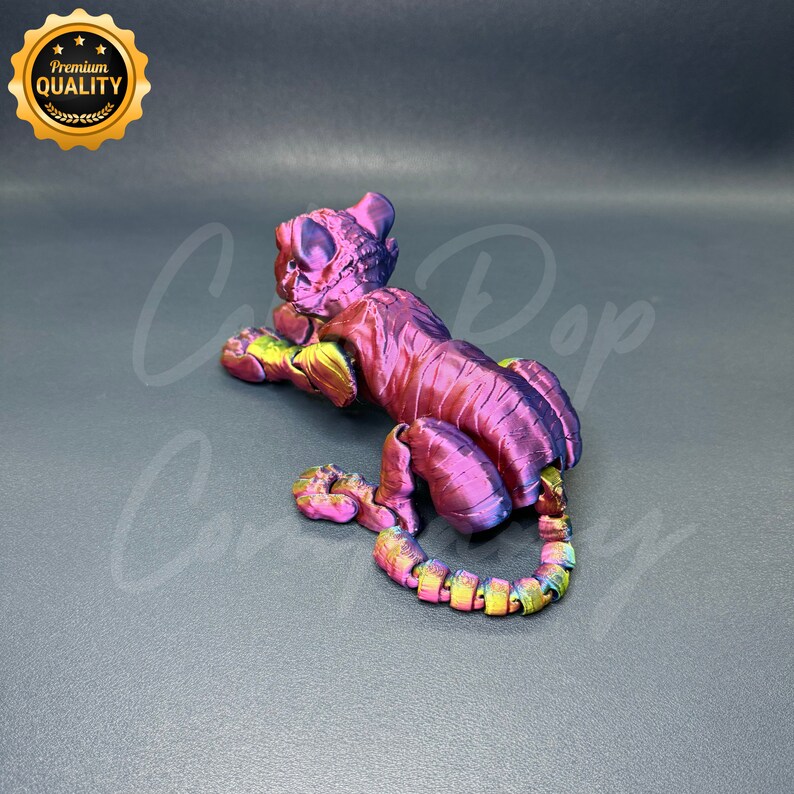 May include: A 3D printed figurine of a tiger in a crouching pose. The figurine is made of a pink and purple iridescent plastic with a metallic sheen. The tiger's tail is curled around its body.