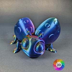 May include: A 3D-printed iridescent bug sculpture, predominantly blue with purple and green hues. It features large, rounded wings and a detailed head. The bug has a textured surface with circular indentations. The sculpture is displayed on a dark grey surface.