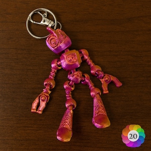 May include: A metallic pink and orange articulated robot keyring. The robot has a head, torso, arms, and legs, all connected with ball joints. The keyring is attached to a silver metal ring.