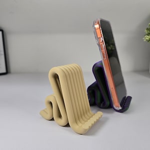 May include: Two modern phone stands. One is beige, with a wavy, ribbed design. The other is purple and green, holding a phone in a clear case. The stands are on a white surface.