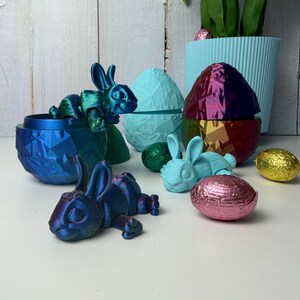 May include: A collection of Easter decorations featuring iridescent blue and purple bunny figurines, geometric egg-shaped containers, and foil-wrapped chocolate eggs. A light blue plant pot is in the background. The items are arranged on a white surface.