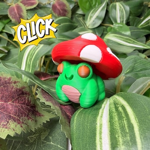 May include: A green frog ornament with orange eyes and a pink belly, wearing a red mushroom hat with white spots. The word "CLICK" is in a yellow speech bubble. The frog is surrounded by green leaves.