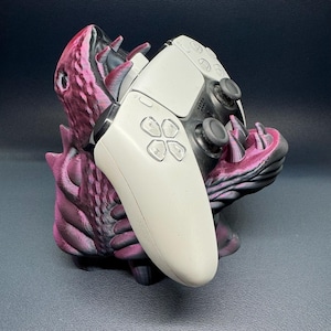 May include: A white and black games controller sits in a purple and pink monster-themed holder. The holder has a textured, scaly appearance with sharp teeth and claws. The controller features a touchpad and button layout.