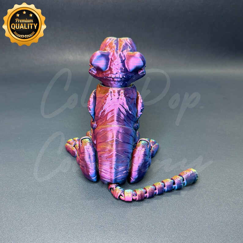 May include: A small, iridescent purple and blue 3D printed figurine of a sitting animal with a segmented tail. The figurine has a textured surface and appears to be a chameleon.