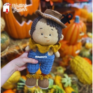 May include: A handmade knitted scarecrow doll with a brown hat, blue overalls, and a yellow collar. The doll has brown hair and is holding a small black crow. The background includes pumpkins and the text "Knitting Pattern".