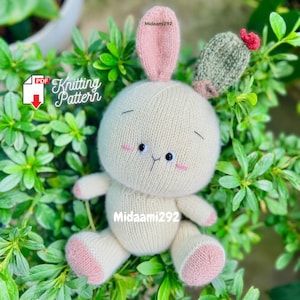 May include: A knitted white bunny rabbit with pink ears and a small green cactus on its head. The bunny has black eyes and a pink nose. The bunny is sitting on a bed of green leaves. The text "Knitting Pattern" is visible in the image.
