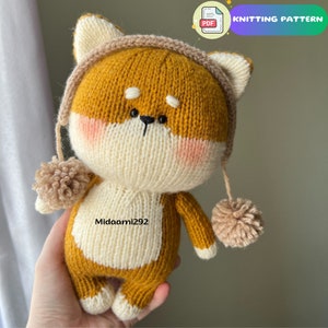 May include: Knitted plush toy dog wearing a brown knitted hat with a pom-pom. The dog is brown and white with black eyes and a black nose. The toy is being held by a hand.