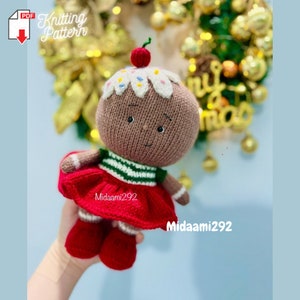 May include: Knitted gingerbread person doll wearing a red dress, green and white striped shirt, and a white icing hat with a red cherry on top. The doll is holding a gingerbread cookie in its hand.