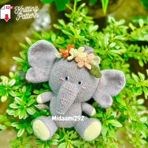 Op de afbeelding: Een gebreid grijs olifantenspeelgoed met een bloemenkroon. De kroon is gemaakt van roze, oranje en gele bloemen. De olifant heeft een kleine, roze neus en zwarte ogen. Het speelgoed zit op een bed van groene bladeren.