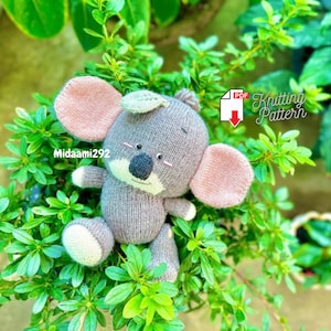 Puede incluir: Juguete de koala de punto gris con orejas rosas y una hoja verde en la cabeza. El juguete está sentado en un lecho de hojas verdes. El texto "Midaami292" y "PDF Knitting Pattern" son visibles en la imagen.