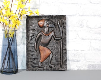 May include: A carved wooden relief of a figure with a staff, in brown and orange tones, against a textured, dark background. A blue glass vase with yellow flowers is in the background.