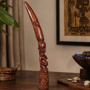 Puede incluir: Una escultura de madera alta tallada a mano que presenta una figura arrodillada en la base, rematada con una extensión larga y curva en forma de cuerno. La madera de color marrón oscuro muestra detalles intrincados y un acabado pulido.