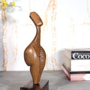 May include: A wooden sculpture with a smooth, curved design, mounted on a rectangular base. The sculpture is a warm brown colour and has a unique, abstract shape. The base is a darker shade of brown. The sculpture is displayed on a grey surface.