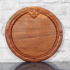 May include: A circular wooden serving tray, featuring a carved face at the top. The tray is a warm brown hue and has a raised edge with decorative carvings. The tray appears handmade, with a rustic, artisanal aesthetic. The diameter is approximately 30 cm.