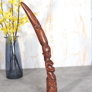Puede incluir: Una escultura de madera alta tallada a mano que presenta una base cilíndrica con patrones geométricos, rematada por una figura humana con rasgos faciales y cabello detallados, que se extiende hacia una punta curva y lisa.