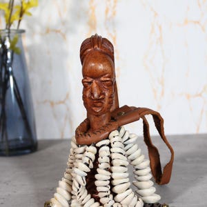 May include: A hand-carved wooden figurine featuring a detailed human face with prominent facial features. The figure is adorned with a brown leather collar and multiple strands of white cowrie shells, accented with small brass bells at the ends.
