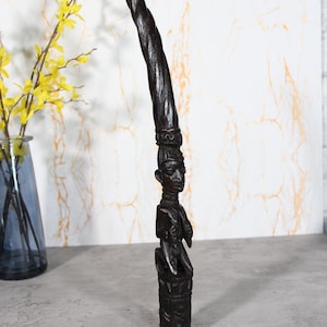 Puede incluir: Una escultura alta de madera tallada a mano en color marrón oscuro, que presenta una figura humana central sobre una base cilíndrica. La figura está rematada por una larga extensión en espiral similar a un cuerno que termina en punta.