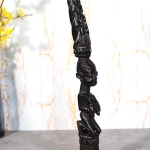 Può includere: Una scultura in legno intagliato marrone scuro di una figura inginocchiata con una testa allungata e un copricapo ornato. La scultura è dettagliata con motivi e texture intagliate, alta circa 30 cm.