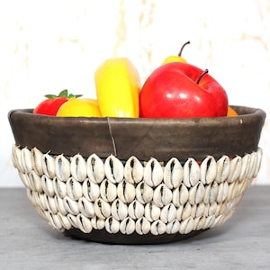 Puede incluir: Un cuenco decorativo con interior marrón oscuro y borde adornado con conchas blancas. El cuenco está lleno de fruta artificial colorida, incluyendo manzanas rojas, pimientos amarillos y un tomate.