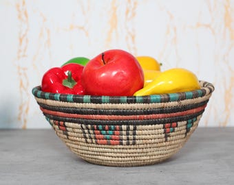 May include: A woven basket filled with colorful, artificial fruit. The basket has a black, red, and turquoise pattern around the rim. The fruit includes a red apple, red bell pepper, green pepper, and yellow lemons.