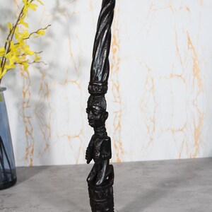 Könnte beinhalten: Eine hohe, dunkelbraune, handgeschnitzte Holzskulptur mit einer Figur auf einem zylindrischen Sockel. Die Figur wird von einem langen, spiralförmig gedrehten Aufsatz gekrönt, der die feine Maserung und die detaillierte Handwerkskunst zeigt.