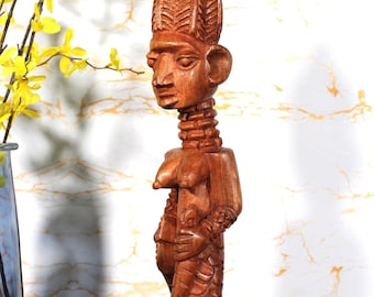 Könnte beinhalten: Geschnitzte Holzstatue einer Figur mit detailliertem Kopfschmuck, langem Hals und stilisiertem Körper. Die Statue hat eine warme braune Farbe und steht auf einem quadratischen Sockel. Die Figur scheint eine kleinere Figur zu halten.