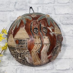 Könnte beinhalten: Runde, braune Wandkunst mit einem geschnitzten Relief, das Figuren mit Gegenständen, Hütten und Palmen darstellt. Das Kunstwerk hat einen dunkelbraunen Hintergrund mit hellbraunen und beigen Details. Ein schwarzer Haken ist zum Aufhängen angebracht.