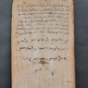 Puede incluir: Una tabla de madera con escritura árabe escrita en tinta negra. El texto dice "Bismillah al-Rahman al-Rahim" y continúa con una serie de versos. La tabla está desgastada y tiene una veta de madera natural.