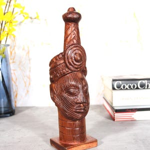 May include: A carved wooden sculpture of a head with an elongated headdress. The sculpture is a reddish-brown color and has detailed facial features and carved patterns. The sculpture is mounted on a square wooden base. The sculpture is a decorative item.