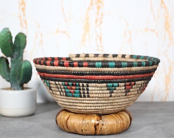 May include: A woven basket with a round shape and a decorative pattern. The basket features bands of red, black, and teal colors. It sits on a round, woven base. A small cactus in a white pot is in the background.