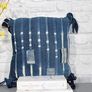 May include: A square throw pillow made of indigo-dyed fabric with vertical white dashed lines and three rectangular patches featuring different striped and grid patterns. The pillow has fabric ties at each corner and rests on a white book.