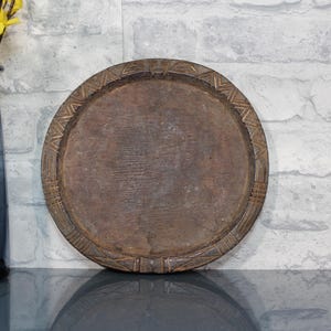 Puede incluir: Una bandeja redonda de madera marrón con detalles tallados alrededor del borde. Se encuentra sobre una superficie reflectante, con un jarrón de cristal azul que contiene flores amarillas a la izquierda. El fondo es una pared de ladrillos blancos.