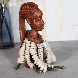 Pode incluir: Um busto de madeira esculpido à mão de uma pessoa com traços faciais detalhados e um penteado alto e texturizado. A figura é adornada com um colar grosso e vários fios de conchas cauri brancas, terminados com pequenos sinos de latão.