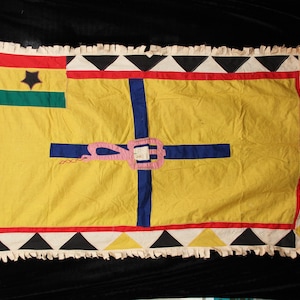 Puede incluir: Una bandera amarilla con una estrella negra sobre una franja roja y verde en la esquina superior izquierda. Una cruz azul y blanca está centrada en la bandera con una cinta rosa y una hebilla. La bandera tiene un borde de flecos blancos y un patrón triangular negro, blanco y rojo a lo largo del borde inferior.