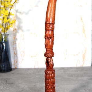 Puede incluir: Escultura de madera tallada a mano, de color marrón rojizo. Presenta un rostro humano estilizado y complejos patrones geométricos. La parte superior tiene forma curva.