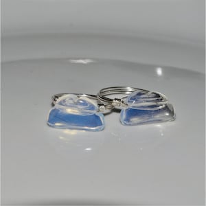 Puede incluir: Dos anillos hechos a mano con una banda de alambre plateado y una piedra transparente azul claro. Las piedras tienen una forma irregular. Los anillos se exhiben sobre una superficie blanca.