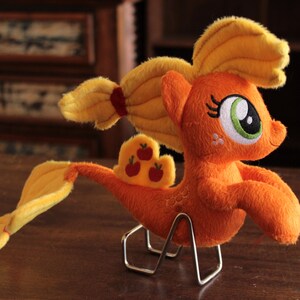Puede incluir: Un peluche naranja con forma de pony con melena y cola amarillas y una cinta roja. El pony tiene un ojo verde y blanco y un pequeño diseño de manzana en el flanco. Se muestra sobre un soporte metálico.