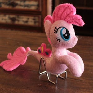 Puede incluir: Un peluche rosa que representa un pony con una gran melena y cola rosas, ojos azules y una mariposa decorativa en la espalda. El juguete se exhibe sobre un soporte metálico, sobre una superficie de madera oscura.