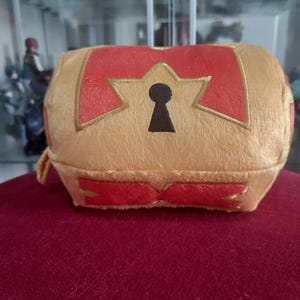Puede incluir: Un cofre de felpa de color dorado con detalles en rojo y un diseño de cerradura. El cofre tiene forma redondeada y está hecho de material suave. Los detalles en rojo están en la parte superior e inferior del cofre.