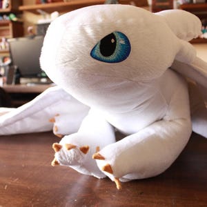 Puede incluir: Un peluche blanco con forma de dragón con grandes ojos azules y pequeñas garras marrones. El dragón tiene grandes alas y está sobre una superficie de madera. El juguete es suave y tierno.