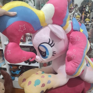 May include: A large plush toy of a pink pony character with a voluminous, multi-colored mane featuring yellow, blue, and pink sections with embroidered white star outlines. The pony has large blue eyes and light yellow hooves decorated with colorful balloon icons.