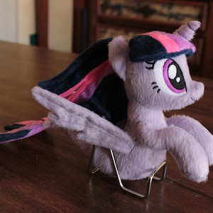 Puede incluir: Un peluche de pony lavanda con melena y cola azul oscuro y rosa. El juguete tiene ojos grandes y se exhibe sobre un soporte metálico. El pony está sobre una superficie de madera.