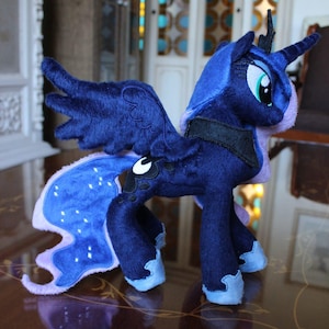 Puede incluir: Un peluche de la Princesa Luna de My Little Pony. El juguete es principalmente azul oscuro con una melena y cola azul claro, y alas grandes y detalladas. Tiene un arnés negro y un cuerno de unicornio. El juguete está sobre una superficie reflectante.