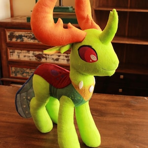 Puede incluir: Un peluche de criatura con cuerpo verde lima, grandes cuernos naranjas y ojos rojos. El juguete tiene una espalda estampada en rojo y verde y un par de alas azules y grises. Está sobre una superficie de madera.
