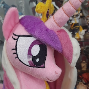 Puede incluir: Un peluche de un personaje de unicornio rosa con crin morada, un cuerno largo y espiral de color rosa, y una corona amarilla con una esfera morada en la parte superior. El personaje tiene grandes ojos bordados con detalles en blanco, morado y negro.