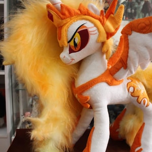 Puede incluir: Un peluche de pony blanco con detalles en naranja y amarillo. El pony tiene una larga melena y cola amarillas, alas naranjas y una corona naranja. Los ojos son una llamativa combinación de amarillo, naranja y rojo.