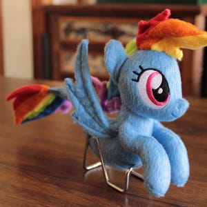 Puede incluir: Un peluche de pony azul claro con melena y cola arcoíris, y grandes ojos rosas y negros. El juguete tiene alas azules y está sobre un pequeño soporte metálico. El pony es un personaje popular de una serie animada para niños.