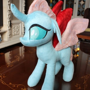 Puede incluir: Un peluche de pony azul claro con ojos turquesa bordados y un cuerno. Tiene alas rosas y rojas. El juguete está sobre una superficie marrón con un diseño floral.