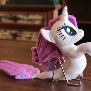 Puede incluir: Un peluche de unicornio blanco con melena y cola moradas, ojos azules y un soporte plateado. El unicornio tiene una cola larga y curvada y descansa sobre una superficie de madera. El juguete está detallado con características bordadas.